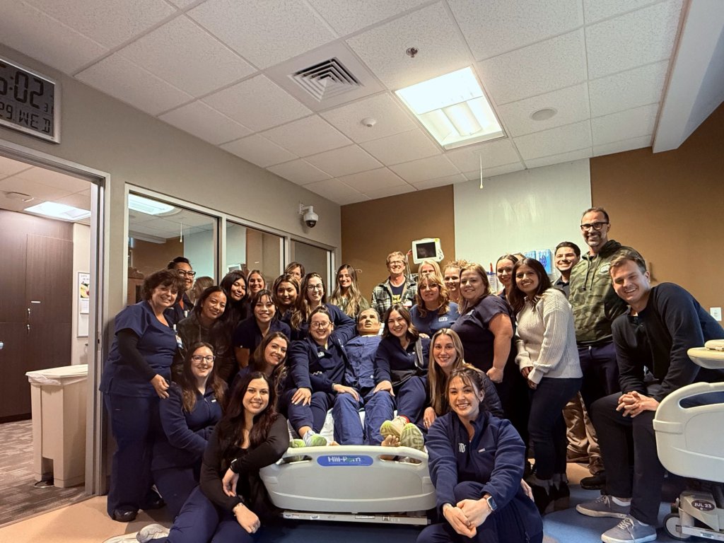 Group photo of ~20 healthcare staff (clinicians, assistants, administrators) in scrubs and business attire, gathered around a dental chair in a modern operatory — smiling for a team celebration or milestone